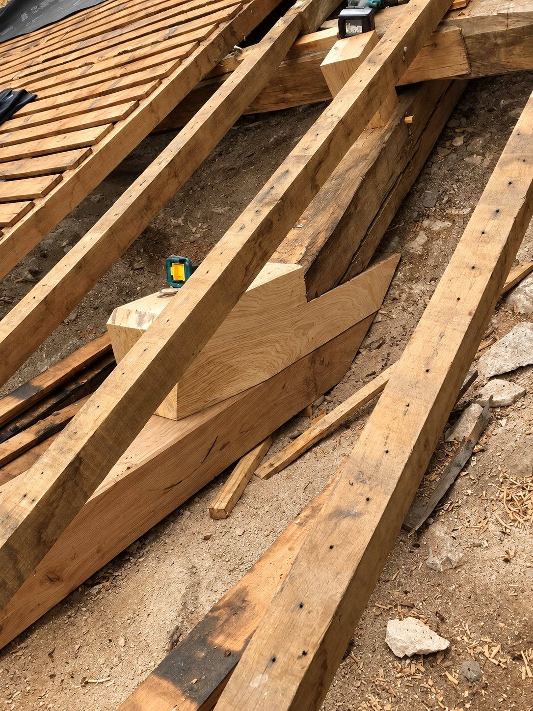 Poutres et pièces de charpente en chêne assemblées sur un chantier, avec un outil posé au centre et de la sciure au sol.