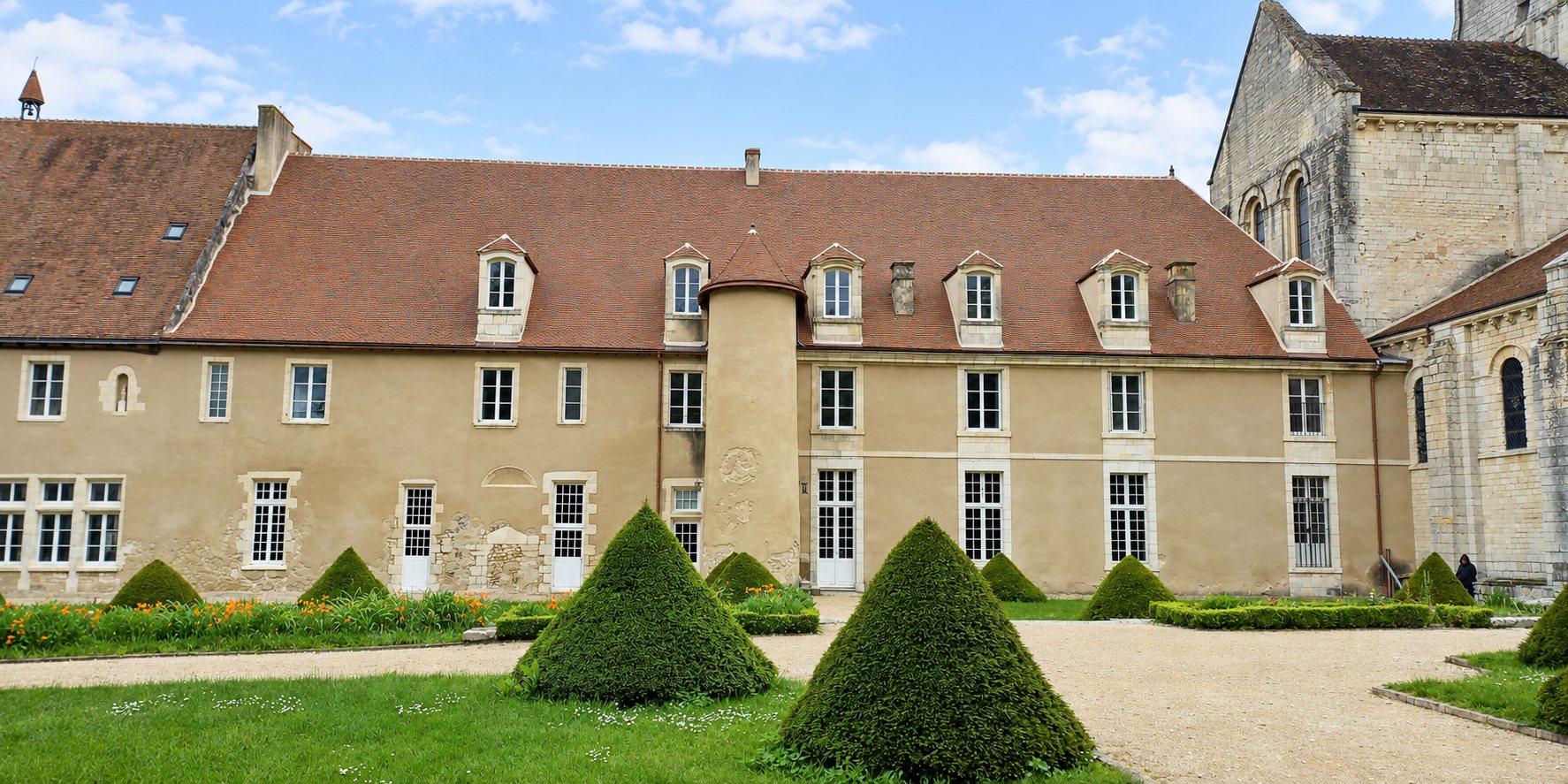 Façade d'un ancien bâtiment en pierre à toit de tuiles rouges, devant un jardin avec des buis taillés en cône et une allée de gravier, à côté d'une église romane.