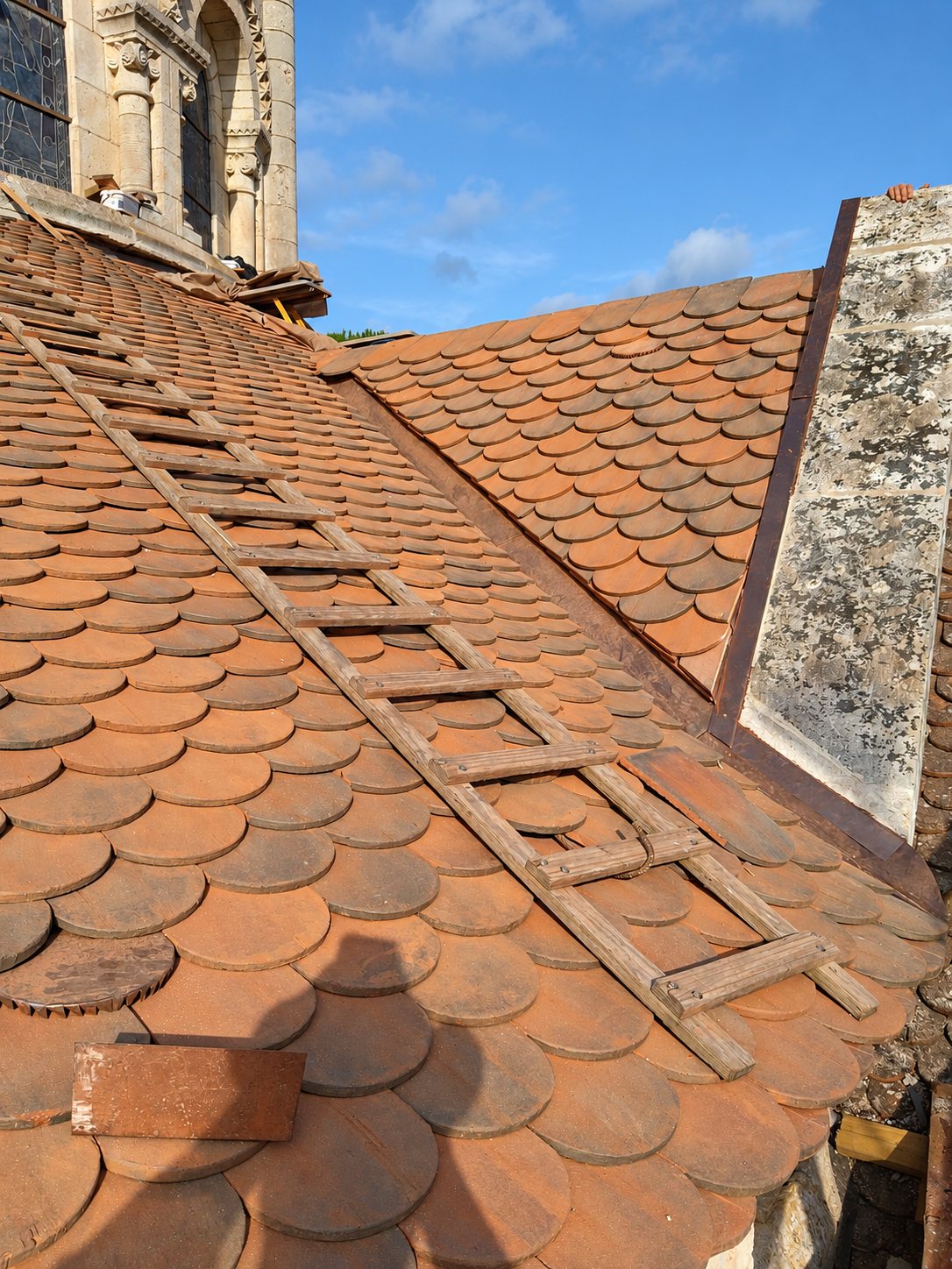 Une échelle en bois est posée sur un toit en tuiles plates de terre cuite, près d'un clocher en pierre sous un ciel bleu.