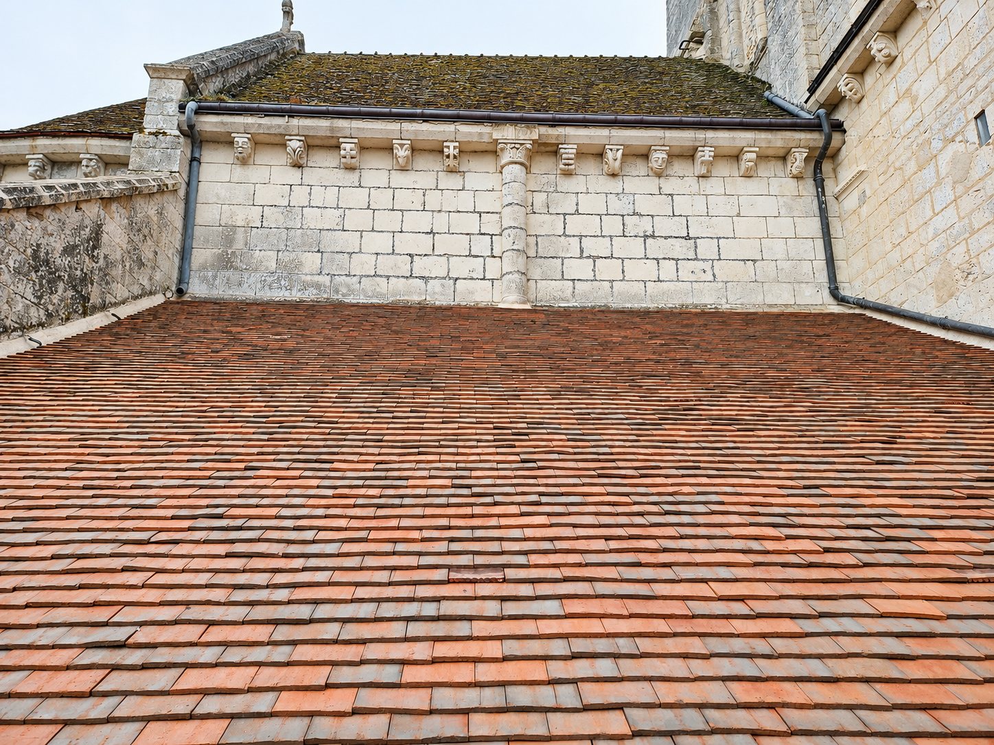 Vue d'un grand toit de tuiles plates rouges et brunes devant un mur en pierre de taille orné de modillons sculptés et d'une colonne.