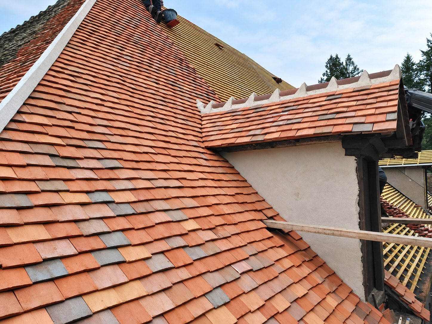 Vue rapprochée d'un toit en tuiles plates de teintes rouges et grises avec une lucarne à rive crénelée, pendant des travaux de rénovation.