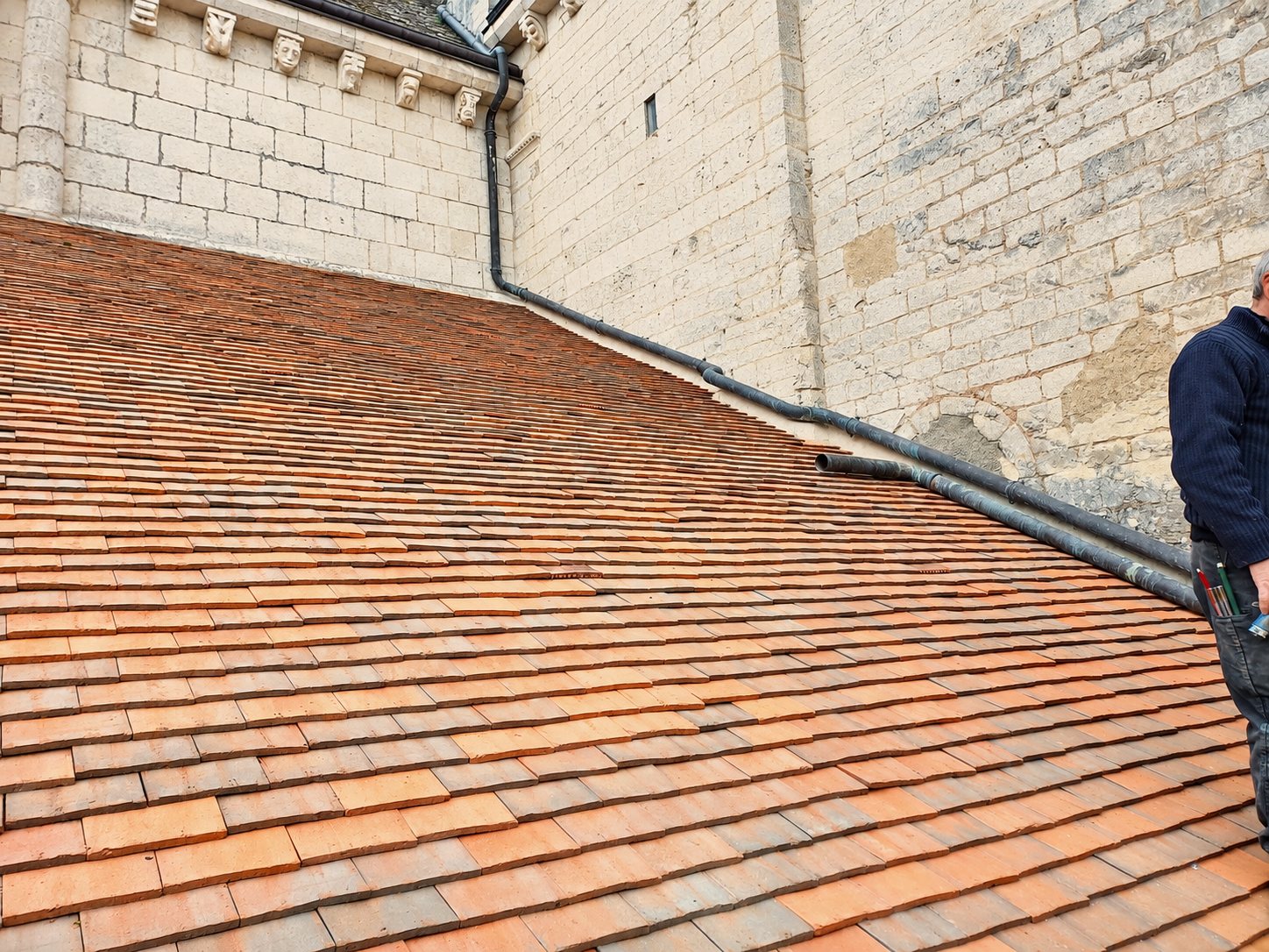 Pente de toit récemment couverte de petites tuiles plates en terre cuite orangée, longée par une descente en zinc, contre un mur de pierre blanche, avec un homme en pull bleu à droite.